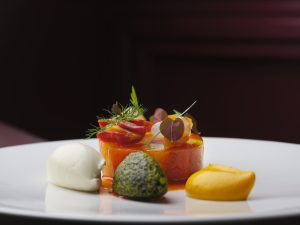 A person in a red blouse holding a white plate with an elegant dessert. The dessert features a layered cake topped with a ribbon-like garnish, accompanied by a scoop of white ice cream on a bed of crumbs. The background is softly blurred.